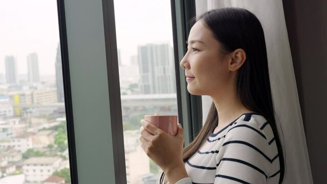 4K Young beautiful Asian woman standing by the window and looking to cityscape with drinking hot coffee or tea in the morning. Happy girl relax and enjoy indoors lifestyle on holiday vacation at home.