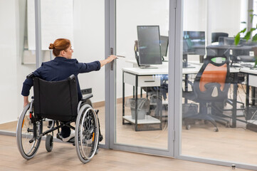 Red-haired caucasian woman in a wheelchair trying to open the door in the office. 