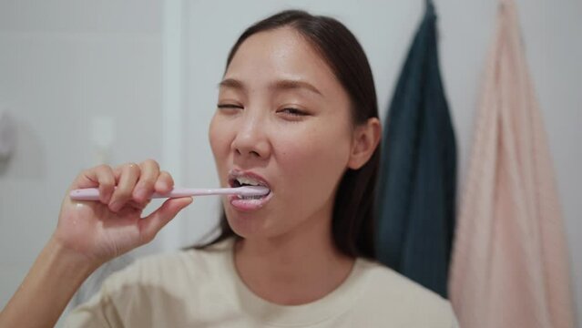 4K Smiling young attractive Asian woman brushing teeth and looking to the mirror in bathroom. Happy girl using toothbrush with whitening toothpaste cleaning teeth in the morning. Dental care concept.