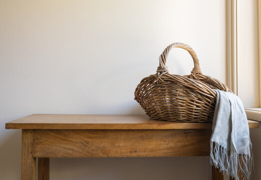 Wicker basket with grey scarf on oak side table next to window against beige wall (selective focus)
