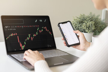 Investor analyzing exchange market, asian young business woman trader on graphic charts on laptop computer, hand in using smartphone for trading stock, looking at screen with diagrams at home office.