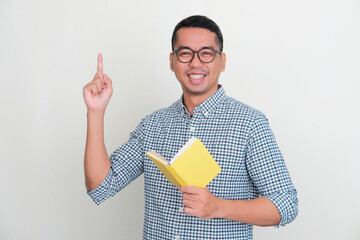 Asian man smiling and pointing finger up while holding a book