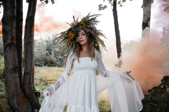 A beautiful young woman in a white sundress and a huge wreath of field herbs poses in a forest clearing, a bride in folk style, boho