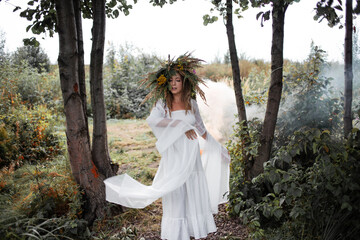 A beautiful young woman in a white sundress and a huge wreath of field herbs poses in a forest clearing, a bride in folk style, boho
