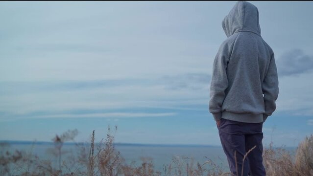A teenage boy stands with his back to the camera on top of a mountain against a clear blue sky at sunset. Ahead of him stretches the sea on the horizon merging with the sky