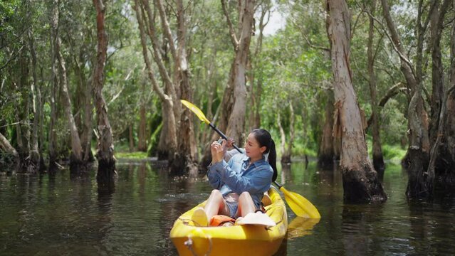 Asian couple kayaking in mangrove forest on summer holiday travel vacation. Man and woman tourist using mobile phone photography beautiful nature while paddling on canoe boat together in forest lake.