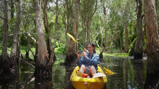 Asian couple kayaking in mangrove forest on summer holiday travel vacation. Man and woman tourist using mobile phone photography beautiful nature while paddling on canoe boat together in forest lake.