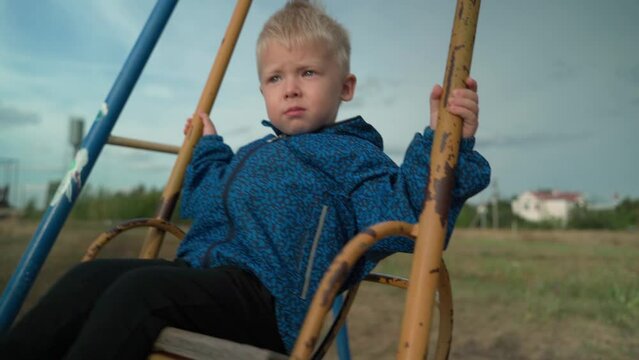 A blond boy sits on a children's swing in the park and slowly swings back and forth. The swing is already very old, which can be seen from the peeling paint on them