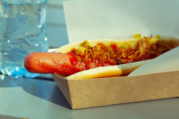 A hot dog and coffee in a plastic cup lie on a white wooden table in close-up. Street fast food in the park or in nature. A quick snack on the go to satisfy your hunger. Eating outdoors on a walk.