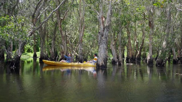 Happy young Asian couple kayaking in mangrove forest on summer travel vacation. Man and woman backpacker tourist paddle on canoe boat together in forest lake. Environment holiday travel trip concept.