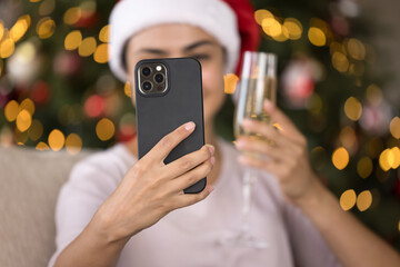 Happy female smartphone user taking selfie with New Year flute of wine at glowing tree in background, making video call for Christmas congratulation, giving wish. Mobile phone in hand close up