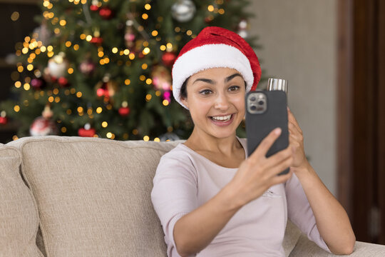 Excited young Indian girl in red Xmas Santa hat taking selfie picture at Christmas tree on smartphone at home, making video call for wishing Happy New Year, smiling at mobile phone webcam, talking
