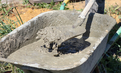 A worker kneads a mixture of concrete in a wheelbarrow at a construction site.