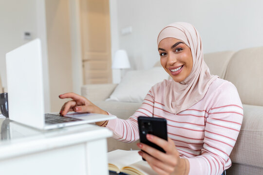 Elegant attractive Muslim woman using mobilephone and laptop searching online shopping information in living room at home. Portrait of happy woman purchasing product via online shopping.