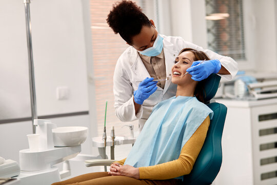 Black Dentist Wearing Face Mask While Examining Teeth Of Young Woman At Dental Clinic.