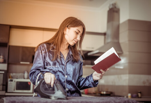Young smiling woman in nightie ironing things with iron and read book in living room