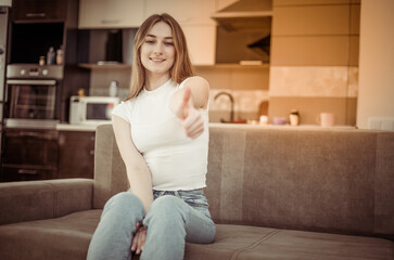 Fototapeta na wymiar Young smiling woman showing thumb up while sitting on sofa in living room