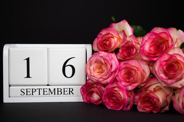 September 16 wooden calendar, white on a black background, pink roses lie nearby.Postcard with copy space. The concept of a holiday, congratulation, invitation, party, announcement, vacation,promotion