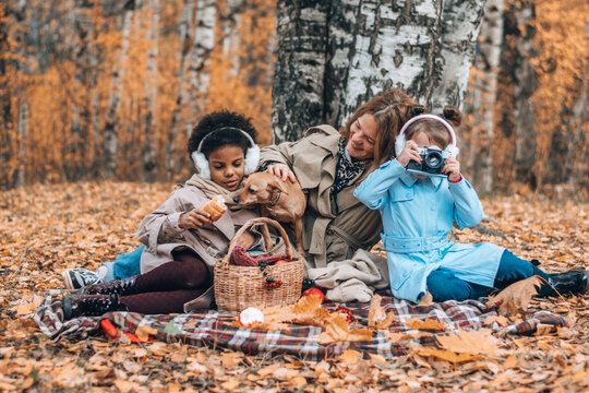 A multiracial family with a dog is having a picnic in the autumn park.Mother and two daughters playing with the dog and having fun.Family,autumn,diverse people concept.