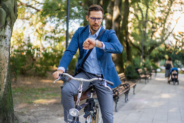 A young business man in a suit is riding a bicycle, he is looking at his wristwatch. The concept of ecological transport