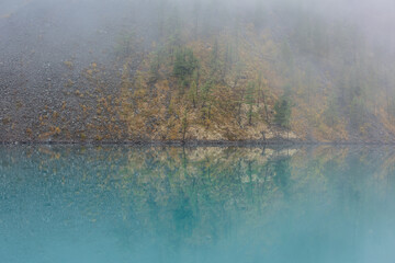 Simple meditative landscape with forest hill reflection in tranquil turquoise lake in fog. Mossy shore with firs in fading autumn colors reflected in mirror mountain lake. Autumn shore of alpine lake.