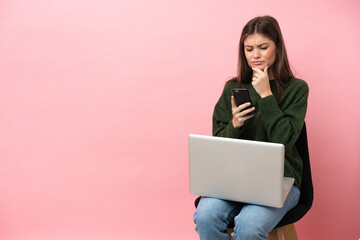 Young caucasian woman sitting on a chair with her laptop isolated on pink background thinking and sending a message