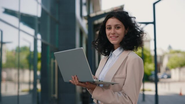 Portrait of the beautiful curly smiling young businesswoman in formal outfit standing near the business centre looking at the camera holding and using laptop for work