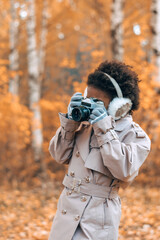 Cute African-American girl in fur headphones takes pictures with a camera in an autumn park.Diversity,autumn concept.