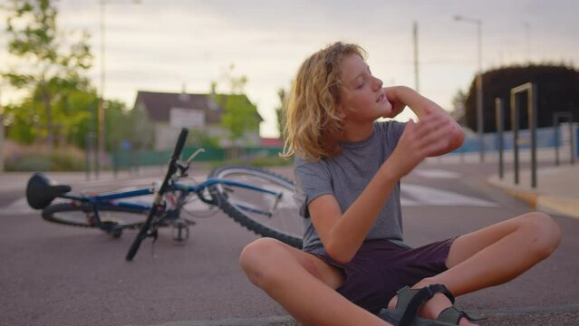 The boy sits on the road after falling off a bicycle, calms the pain in his elbow. Broken bike in the background