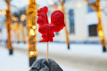 Hand in mitten holding rooster-shaped lollipop on a Christmas market