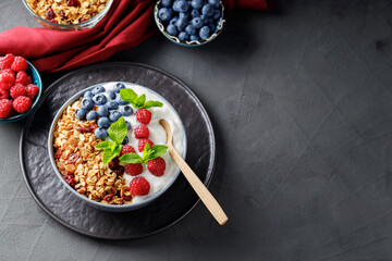 Breakfast cereal granola with berries and greek yogurt on a dark background. Bowl with yogurt, granola and fresh berries on a black plate. Healthy breakfast with wooden spoon