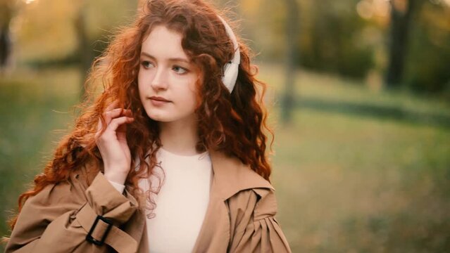 Gorgeous young red head woman listening to music in headphones standing in the park. Portrait of beautiful ginger girl.