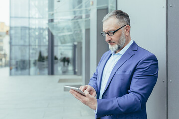 Senior handsome businessman man in glasses and a suit stands on the street near the office center and waits for customers, employees, holds the phone in his hands, dials a number, messages.