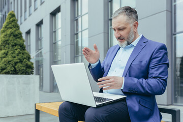 Fototapeta premium Successful senior male businessman conducts a video conference from a laptop. Sitting outside in a suit on a bench near an office center, waves his hands, explains.