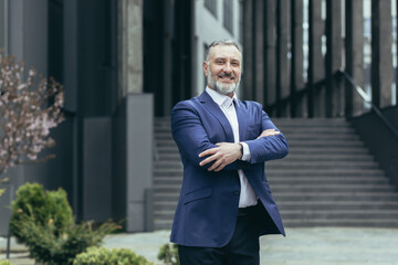 Portrait of a successful senior male businessman in a business suit standing confidently near the office center in full height, arms crossed, smiling. © Liubomir