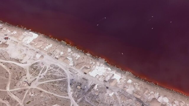 Aerial footage of stacks of salt and roads made of salt along a pink lake, clouds reflecting on the lake, Retba lake, Dakar, Senegal, facing down at the shore and going towards it while tilting down.