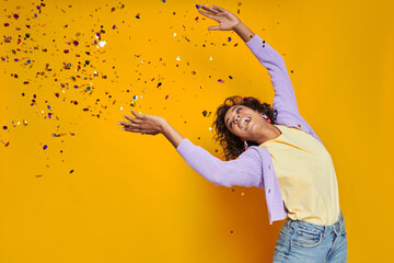 Fototapeta na wymiar Happy African woman throwing confetti and smiling while standing against yellow background