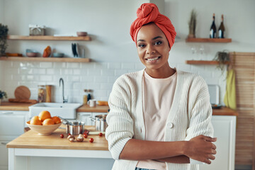 Cheerful African woman in traditional headwear looking at camera while standing at the kitchen