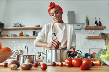 Confident African woman mixing something in pan and smiling while preparing food at the kitchen