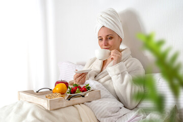 Woman eating fruit breakfast in bed at home in the morning. Lady wrapping a towel around head. Girl wearing bathrobe on clean white bedding with cozy blanket. Healthy lifestyle