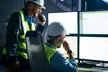 Main engineer and workers operator wearing safety vests and hard hats control product process on factory uses SCADA system and industry 4.0. Two operators follow assembly line using screens with UI