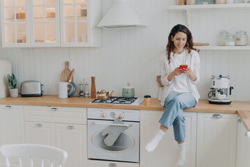 Female housewife satisfied with house cleaning services sits in modern clean kitchen with smartphone