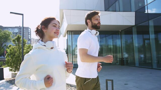Side view of happy caucasian couple with headphones on neck exercising and training on sunny day along glass wall of business center. Concept of sport and healthy lifestyles.