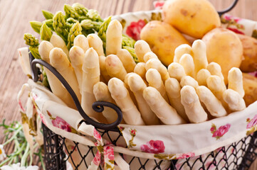 Raw green and white asparagus with potatoes in a black basket offered as close-up on a rustic wooden board
