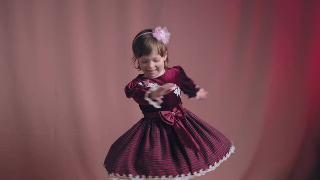 Cute little girl in an vintage burgundy dress is dancing, rejoicing and making funny faces. The concept of children theater productions and developing acting circles. Pink background