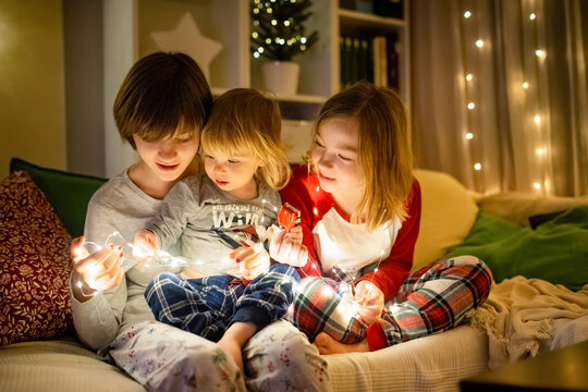 Two big sisters and their toddler brother playing with Chistmas lights in a cozy living room on Christmas eve. Kids spending time at home during winter break.