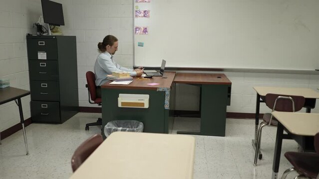 Lonely teacher sitting at desk in empty classroom grading papers.