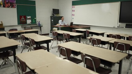 Lonely teacher sitting at desk in empty classroom grading papers.