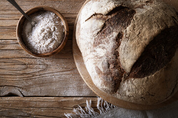 Long leavening wholemeal bread, cutting board and bowl with flour on wooden background, top view,...