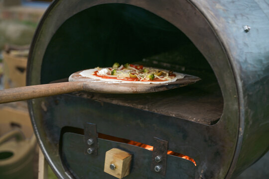 Fresh Pizza On A Wooden Slide Is Pushed Into A Self-made Recycling Oven Built From An Old Metal Barrel, Creative Upcycling Concept, Copy Space, Selected Focus